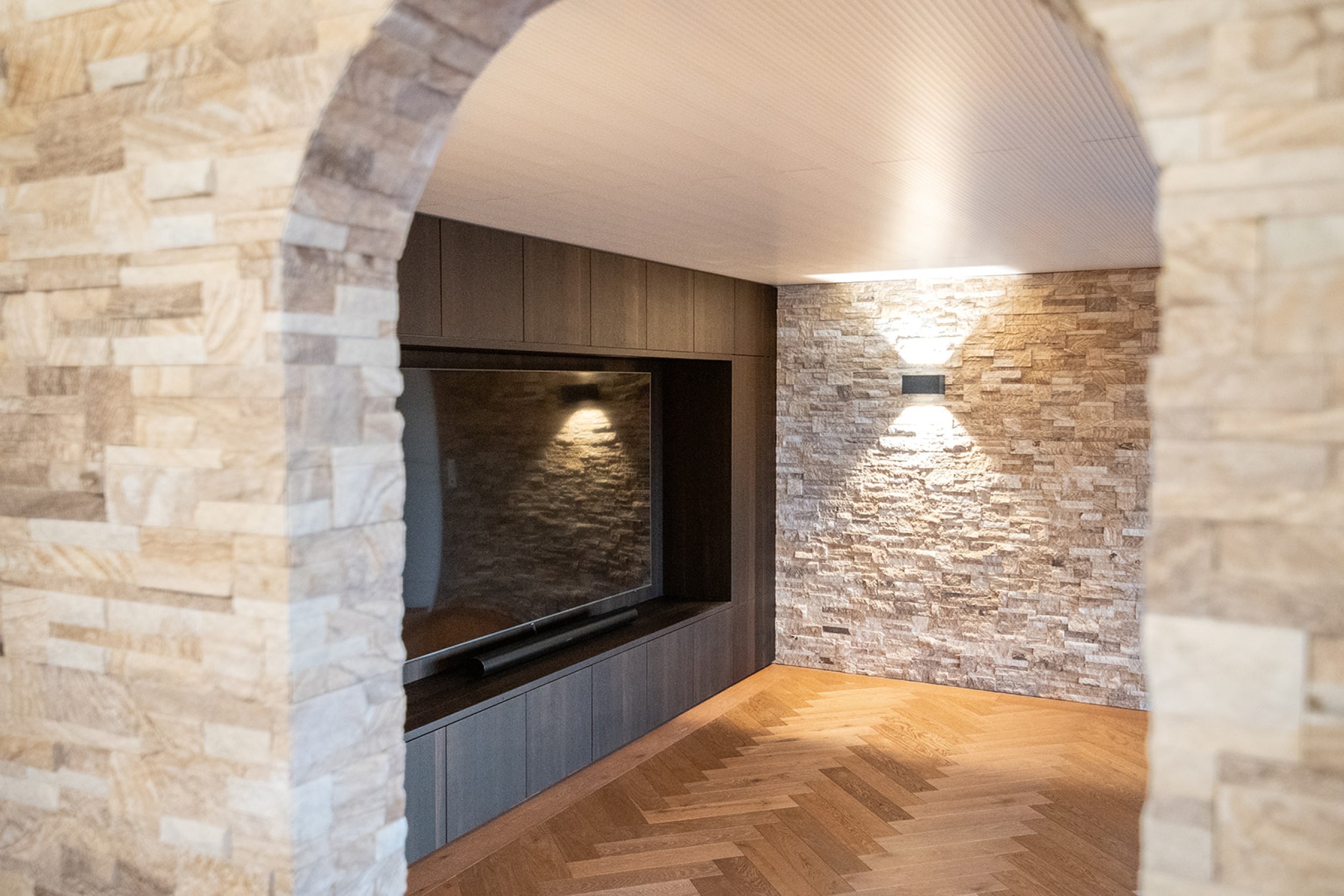 Modernes Untergeschoss mit Home Cinema und Bar, geprägt von rustikalem Steinmauerwerk und elegantem Fischgrat‑Parkett.